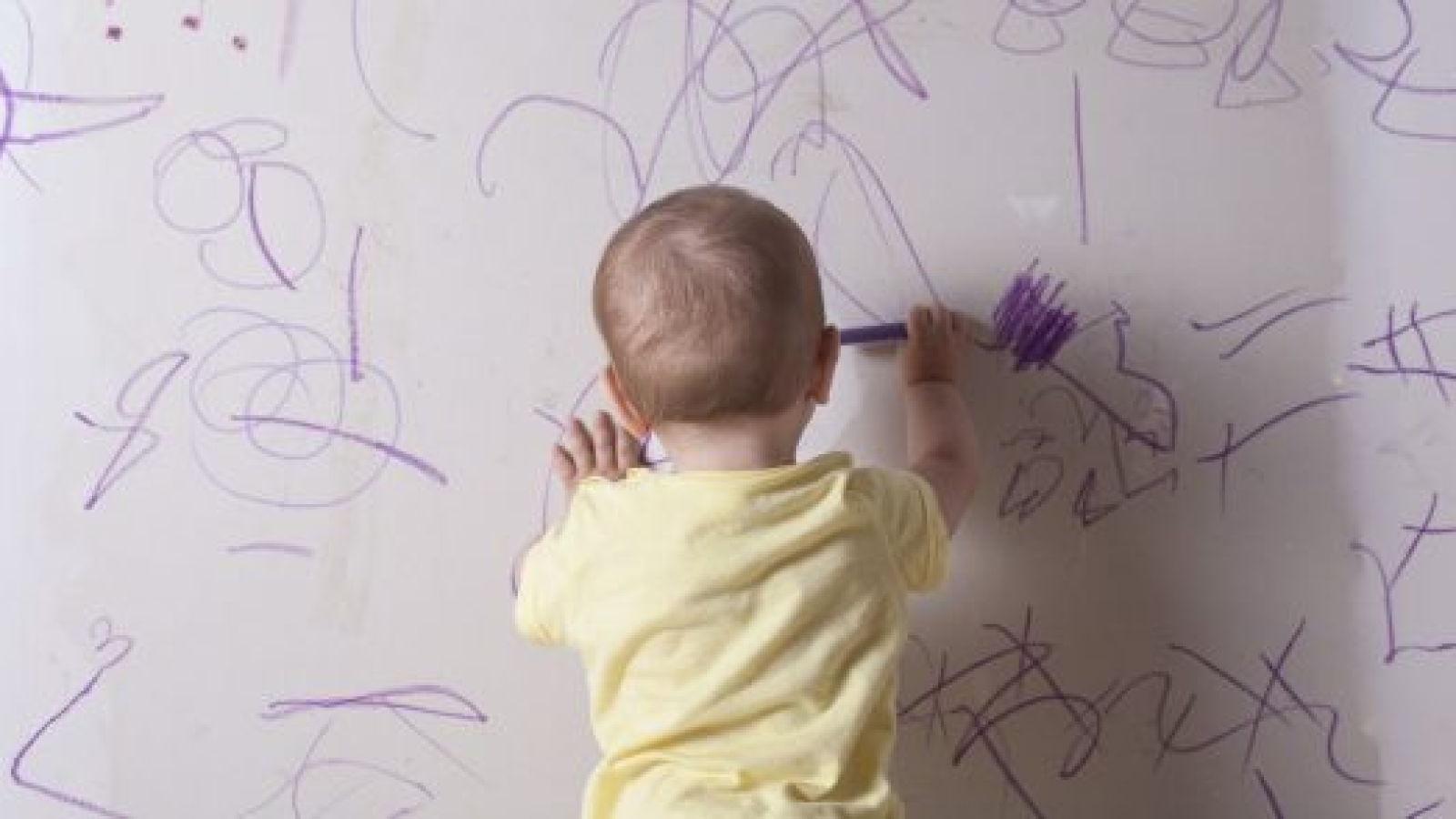 Baby boy drawing with wax crayon on plasterboard wall. He is with his back towards