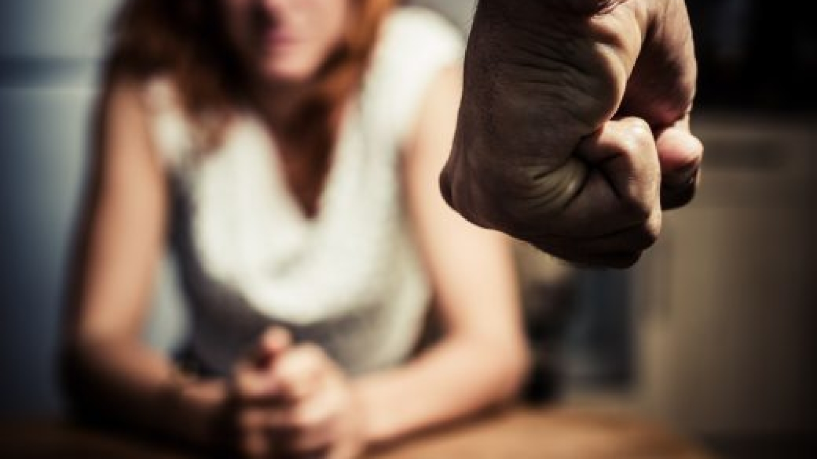Young woman is sitting hunched at a table at home, the focus is on a man's fist in the foregound of the image