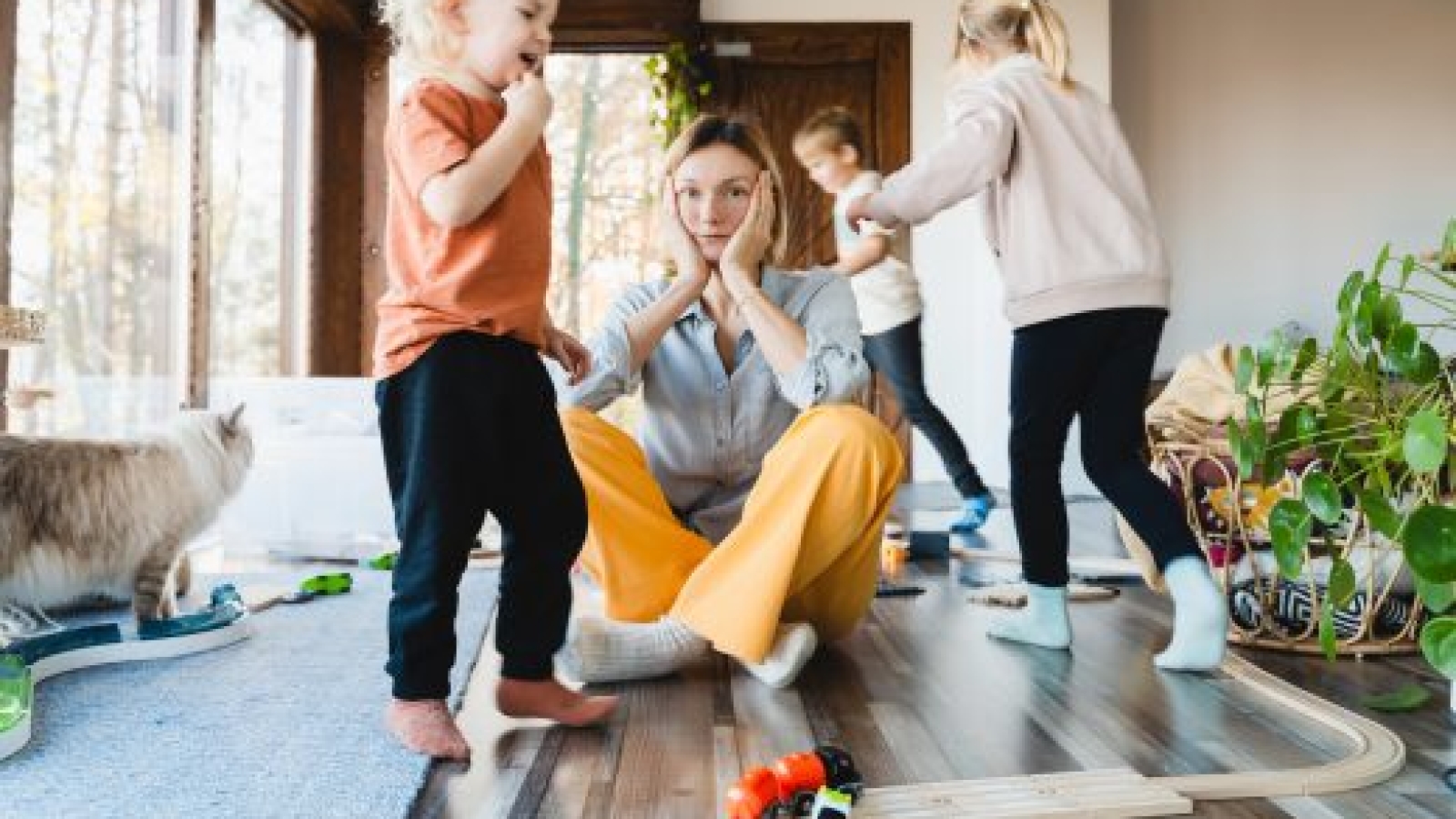 Frustrating woman alone struggling burnout with kids. Stressed out mother sitting on floor in middle of toys while children naughty running around her at room. Family home with chaos, mess.