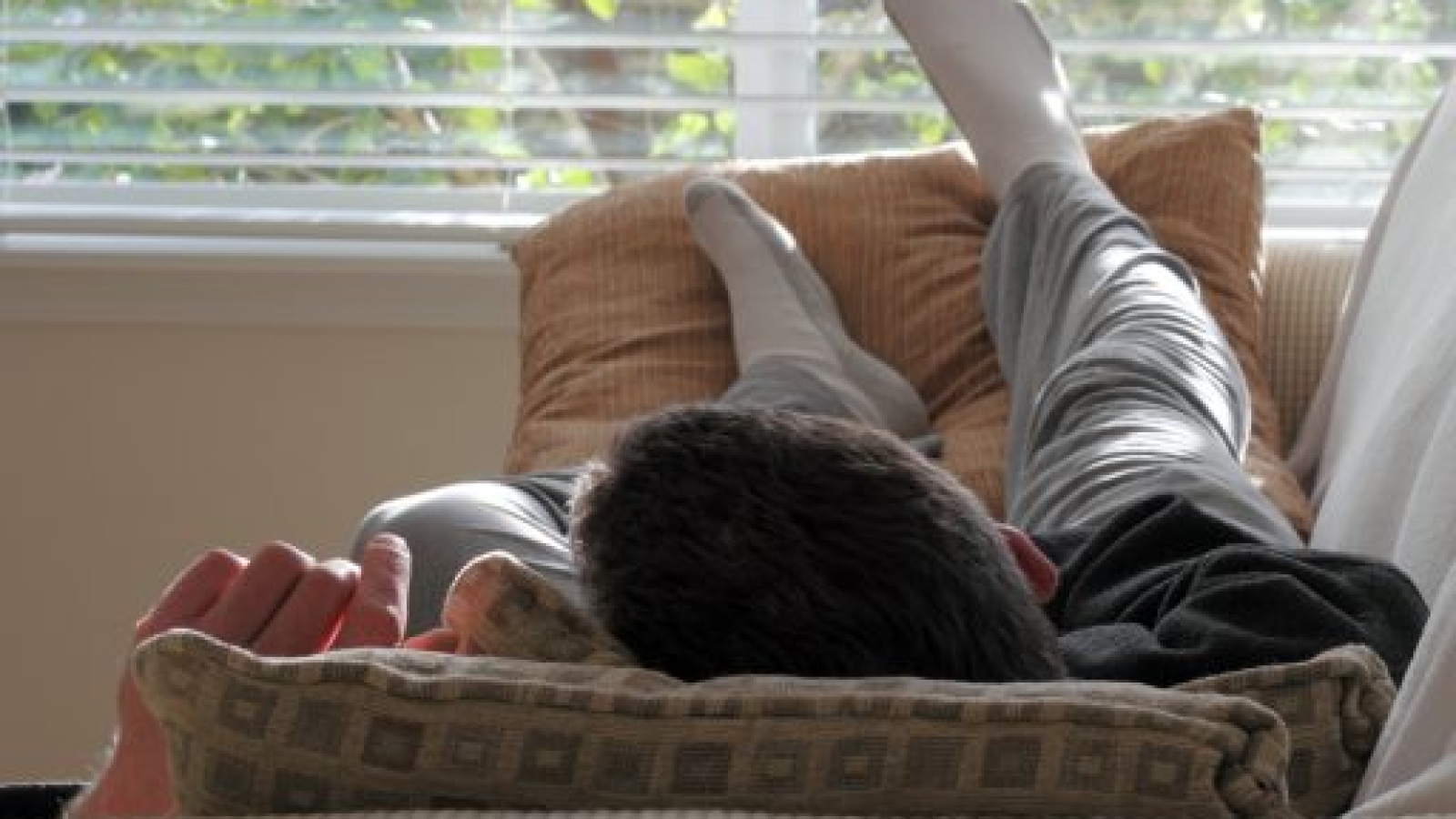 Adult caucasian male laying with his head on a pillow on a couch with his feet up on some pillows in front of a bright living room window in the afternoon..